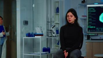 Woman waiting doctor sitting on chair in neurological research laboratory, while researchers team discussing in background health status of patient, brain functions, nervous system, tomography scan video