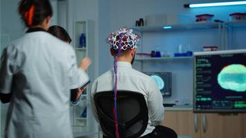 Back view of man patient wearing performant brainwave scanning headset sitting in neurological research laboratory while medical researcher adjusting it, examining nervous system typing on tablet. video