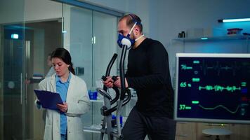 Monitor shows EKG reading of man athlete running on cross trainer and discussing with medical reseacher. Specialist supervising exercise process, controlling physical activity and muscular endurance video