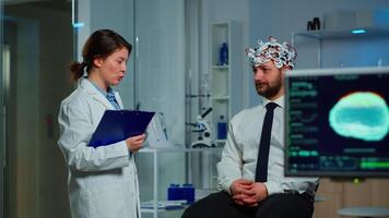Man sitting on neurological chair with brainwave scanning headset while researcher examining health status writing on clipboard. Doctors working in brain study lab with monitors showing EEG reading. video