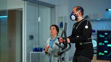 Medical researcher talking with sportsman and showing EKG scan on virtual screen while measuring his endurance using body sensors, electrodes and mask monitoring cardiac rhythm in sports science lab video