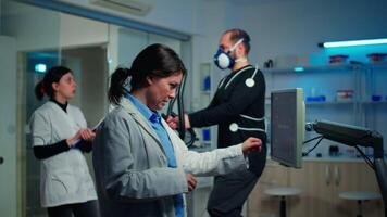 Doctor sport scientist checking EKG scan at monitor and discussing while man with mask running on cross trainer monitoring his endurance. Team of researchers monitoring his endurance and heart rate video