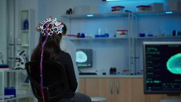 Back view of woman patient wearing performant eeg headset sitting on chair in neurological research laboratory while medical researcher adjusting it, examining nervous system typing on tablet. video