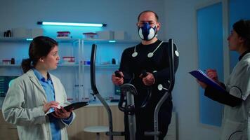 Athlete with mask and electrodes attached on body training with cross trainer while sport scientist interacs with tablet and supervises EKG status. Team of researchers using high-tech equipment video