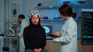 Stressed patient sitting on neurological chair with eeg headset, researcher examining health status writing on tablet. Medical scientist looking through microscope in background developing treatment. video