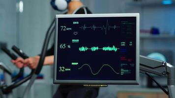 Monitor showing EKG scan of athlete running on cross trainer and medical specialists supervise exercise in background, controlling physical activity, measuring heart rate in sports science laboratory video
