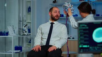 Woman researcher putting brainwave scanning headset to patient developing experiments using brain study lab monitors. Man siting on chair while scientist adjusting device examining nervous system video