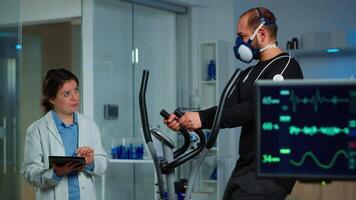 Monitor showing EKG reading of athlete running on cross trainer, while medical specialists supervise exercise, controlling physical activity, measuring heart rate in sports science laboratory video