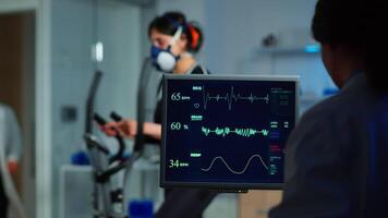 Medical researcher examining EKG image showing on monitor while patinet with mask running on cross trainer testing heart rate using electrodes.Doctor monitoring physical endurance in science sport lab video