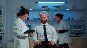 Young man wearing brainwave scanning headset discussing with neurological researcher about symptoms of disease. Medical scientist writing informations of brain scan on tablet working in laboratory video