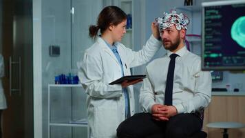 Man sitting on neurological chair with brainwave scanning headset, researcher doctor tracking evolution of patient's brain using tablet, adjusting eeg device. Scientist examining nervous system. video