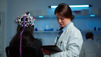 Woman patient wearing performant eeg headset scanning brain electrical activity in neurological research laboratory while medical researcher adjusting it, examining nervous system typing on tablet. video