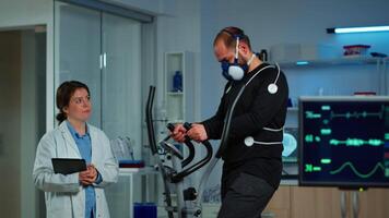 Patient making electrocardiogram during physical exercise in sports science lab, monitoring endurance and heart rate, computer showing EKG data on display. Man running with mask and medical electrodes video