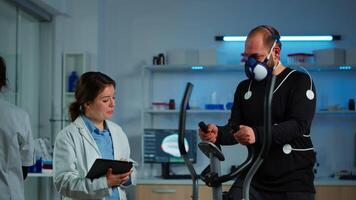 Medical researcher talking with sportsman while measuring his endurance using body sensors, electrodes and mask monitoring cardiac rhythm. Patient running on cross trainer in sports science lab video