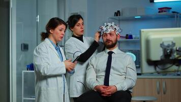 Neurological researchers explaning treatment result pointing on monitor while medical scientist adjusting brainwave scanning headset preparing for brain scan analysing electrical activity video
