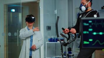 Scientist doctor in sport science laboratory wearing virtual reality goggles while athlete running with electrodes attached on body monitoring physical endurancewhile ekg scan runs on computer screen. video