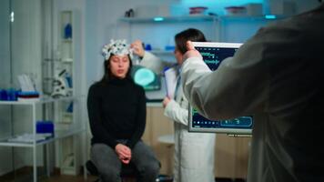Stressed patient sitting on neurological chair with eeg headset, testing health status of brain, while medical researcher looking at monitor with tomography scan developing treatment in laboratory video