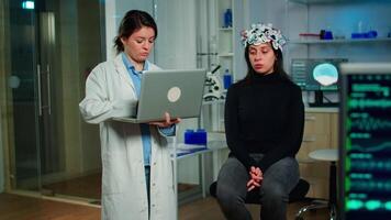 Medical neurologist doctor asking symptoms of patient taking notes on laptop, woman wearing high tech brain scanner. Researcher scientist controlling EEG headset analyzing nervous system functions video