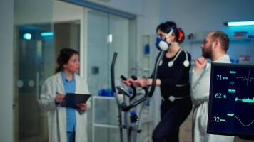 Sportswoman running with mask on cross trainer with electrodes attached to body. Physician using tablet computer and controls EKG data showing on laboratory monitors, discussing with patient video