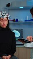 Vertical . Stressed patient sitting on neurological chair with eeg headset, researcher examining health status writing on tablet. Medical scientist looking through microscope in background developing treatment video