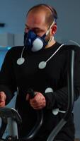 Vertical . Doctor science typing on tablet while measuring sportsman endurance using body sensors and mask monitoring cardiac rhythm. Man running on cross trainer in sports lab, specialists supervise exercise video