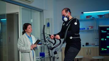 Slow motion shot of sport scientist looking at tablet with EKG scan while man with mask running on cross trainer monitoring his endurance. Team of researchers monitoring his endurance and heart rate video