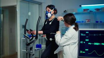 Woman athlete with mask running on cross trainer after medical researcher attached electrodes to her body. Physician using computer to controls EKG data showing on laboratory monitors, VO2 max test video