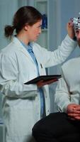Vertical . Man sitting on neurological chair with brainwave scanning headset, researcher doctor tracking evolution of patient's brain using tablet, adjusting eeg device. Scientist examining nervous system. video