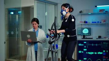 Doctor scientist holding laptop computer and controling EKG data showing on laboratory monitors. Lab science researcher measuring endurance of athlete, heart rate using electrodes and mask video