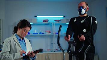 Man athlete running on cross trainer with electrodes attached to his body and mask. Physician using tablet computer and controls EKG data showing on laboratory monitors, discussing with patient video