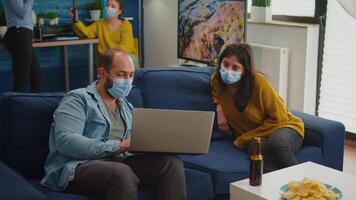 Man with mask showing new movie on laptop to woman sitting in living room keeping social distancing against coronavirus pandemic prevent virus spread. People socializing during covid 19 outbreak video