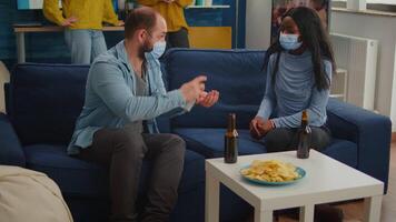 Group of multi ethnic using hand sanitizer gel to prevent covid19 spread spending time together in living room sitting on sofa wearing face mask. Diverse people socializing keeping social distance video