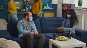 Diverse friends discussing during new normal party wearing protection mask, socializing keeping distance sitting on sofa drinking beer and eating snaks. Group of multi ethnic people enjoying free time video