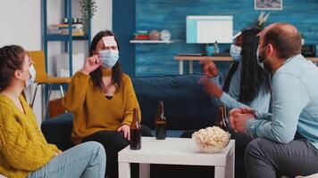Diverse group of funny people in living room playing Who am I game with sticky papers attached to foreheads during global pandemic wearing protection masks. Friends socializing in outbreak video