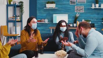 Group of multiracial colleagues with protective masks playing game with sticky notes. Black woman trying to guess what is written on forehead keeping social distancing preventing covid19 spread video