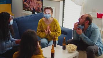 Funny friends with sticky notes on foreheads enjoying name game gesturing during new normal party wearing face mask keeping social distancing preventing covid19 spread. People enjoying bottles of beer video