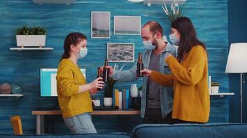 Friends wearing face protective medical masks for protection from virus disease drinking beer, clinking bottles having fun during new normal party in living room. People socializing during outbreak video