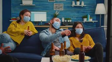 Diverse people relaxing together watching tv holding remote control keeping social distancing wearing mask during coronavirus outbreak to prevent illness. Enjoying party drinking beer and eating chips video
