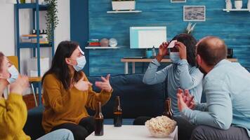 Thoughtful multiethnic friends with sticky notes on foreheads playing name game wearing face mask keeping social distancing preventing covid19 spread drinking beer enjoying popcorn. Conceptual image. video
