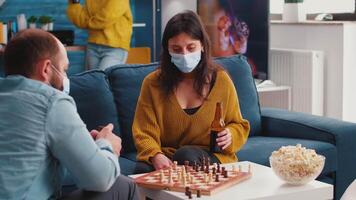 Focused woman wearing face mask playing chess with man drinking beer, keeping social distancing preventing spread of covid 19 virus. Diverse people having fun in home living room playing board games video