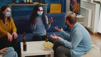 Thoughtful multiethnic friends with sticky notes on foreheads playing name game miming gesturing wearing face mask keeping social distancing preventing covid19 spread. People enjoying bottles of beer video