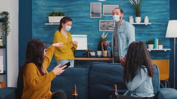 Young people from diverse cultures taking selfie wearing protection masks having fun together during new normal party keeping social distancing, holding bottles of beer enjoying time in living room video