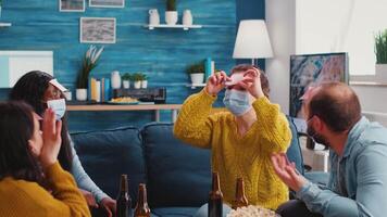 Mixed races friends having fun playing game with sticky notes on foreheads, drinking beer, enjoying new normal party. People wearing face mask keeping social distancing preventing covid19 spread video