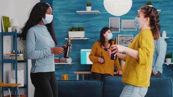 Happy diverse friends meeting at new normal party greeting each other touching elbow holding bottles of beer, having fun. Wearing face mask preventing spread of covid 19 virus enjoying free time video
