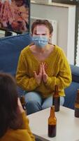 Vertical . Funny friends with sticky notes on foreheads enjoying name game gesturing during new normal party wearing face mask keeping social distancing preventing covid19 spread. People enjoying bottles of beer video