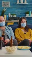 Vertical . Diverse group of friends watching TV together comedy sitcom eating snacks and drinking beer, clinking bottles and having fun at new normal party, wearing protection face mask against covid 19 virus video
