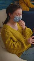 Vertical . Young relaxed woman taking off face mask looking at camera drinking beer sitting on couch while speding time with friends during global pandemic with coronavirus. Group of young people having fun. video