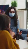 Vertical . African woman playing name game with sticky notes on forehead having fun with friends during new normal party. Wearing face mask in living room keeping social distancing to prevent spreading virus video