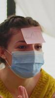 Vertical . Happy woman gesturing playing name game with multiethnic friends having sticky notes on forehead wearing face mask keeping social distancing. Diverse group of people having fun at new normal party video