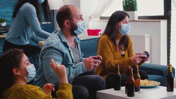 Excited diverse friends winning games using joystick having fun wearing face mask to prevent spreading of coronavirus in time of global outbreak. Gaming competition enjoying beer and popcorn. video
